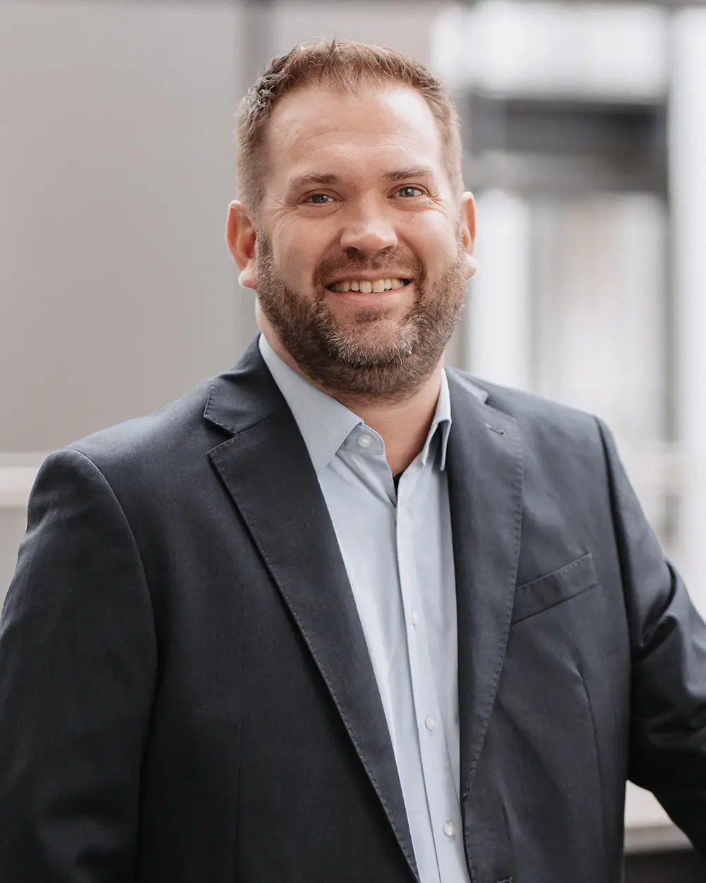 guido_karte Ein Mann mit kurzen Haaren und Bart, der eine dunkle Anzugsjacke und ein hellblaues Hemd trägt, steht in einem Haus und lächelt in die Kamera.