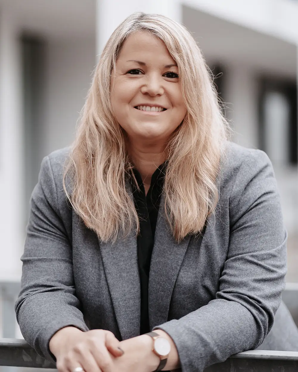 verena_karte Eine Frau mit langen blonden Haaren, die einen grauen Blazer und ein schwarzes Hemd trägt, lehnt sich lächelnd an ein Geländer im Freien.
