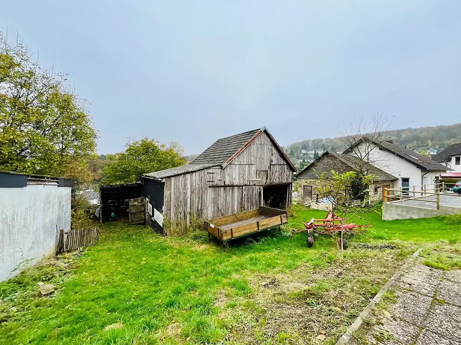 Objekt 1 Kopie Eine verwitterte Holzscheune mit einem Anhänger und alten landwirtschaftlichen Geräten steht auf einem grasbewachsenen Grundstück, mit Häusern und Bäumen im Hintergrund unter einem bewölkten Himmel.