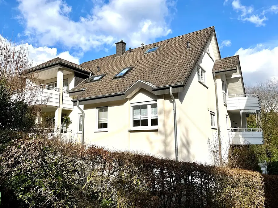 Objekt 10 Ein zweistöckiges, cremefarbenes Haus mit einem schrägen braunen Dach, mehreren Balkonen und umliegenden Hecken, unter einem teilweise bewölkten Himmel.