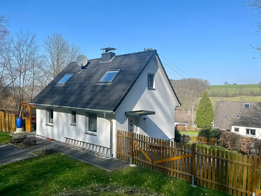 Objekt 11 Ein kleines weißes Haus mit einem dunklen Schrägdach, Oberlichtern und einer Satellitenschüssel, umgeben von einem Holzzaun und einem grasbewachsenen Hof unter einem klaren blauen Himmel.