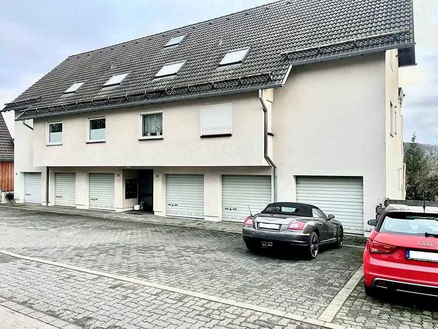 Objekt 12 Ein zweistöckiges Gebäude mit fünf geschlossenen Garagentoren im Erdgeschoss; davor sind zwei Autos geparkt, ein schwarzes Cabrio und eine rote Limousine.