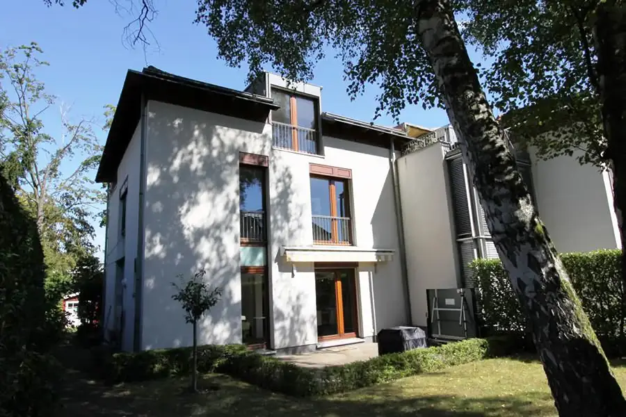 Objekt 15 Modernes dreistöckiges weißes Haus mit großen Fenstern, einem kleinen Balkon und einem Innenhof, umgeben von Grün und Bäumen an einem sonnigen Tag.