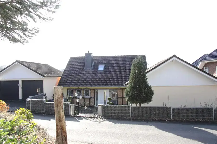 Objekt 16 Einstöckiges Haus mit dunklem Schrägdach, kleinem Vorgarten und angebauter Garage, umgeben von einem niedrigen Steinzaun in einer ruhigen Wohnstraße.