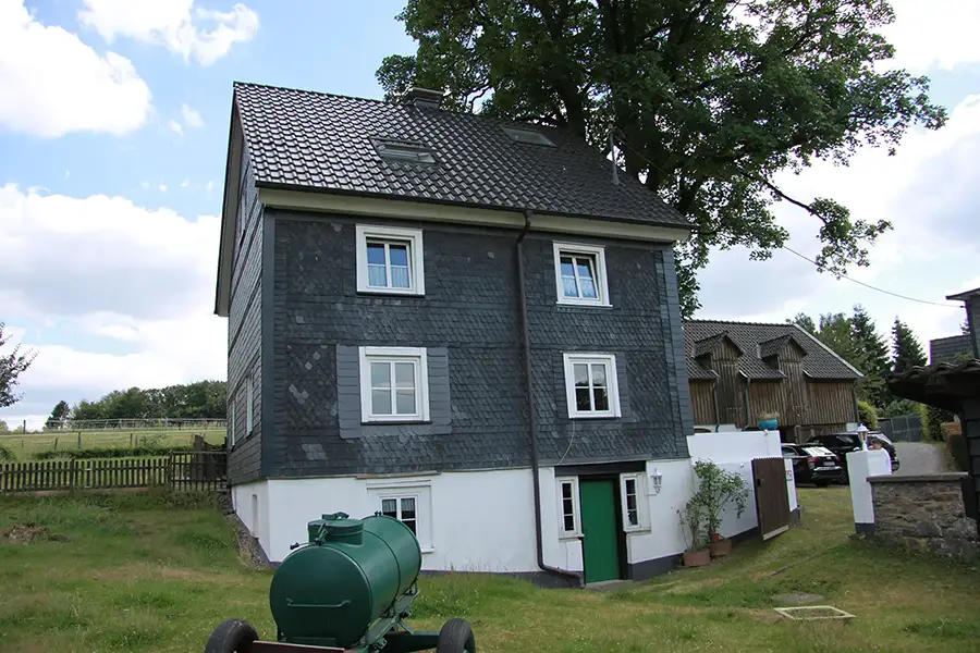 Objekt 17 Ein dreistöckiges Haus mit dunkler Schieferverkleidung, weiß gerahmten Fenstern und einer grünen Tür, das auf einem grasbewachsenen Grundstück mit Bäumen und einem Wassertank im Vordergrund steht.