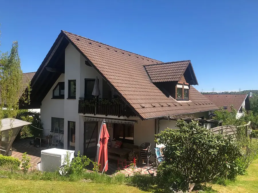 Objekt 18 Ein zweistöckiges Haus mit einem braunen Schrägdach, weißen Wänden, einem Balkon und einer Terrasse mit Gartenmöbeln, umgeben von Grün unter einem klaren blauen Himmel.