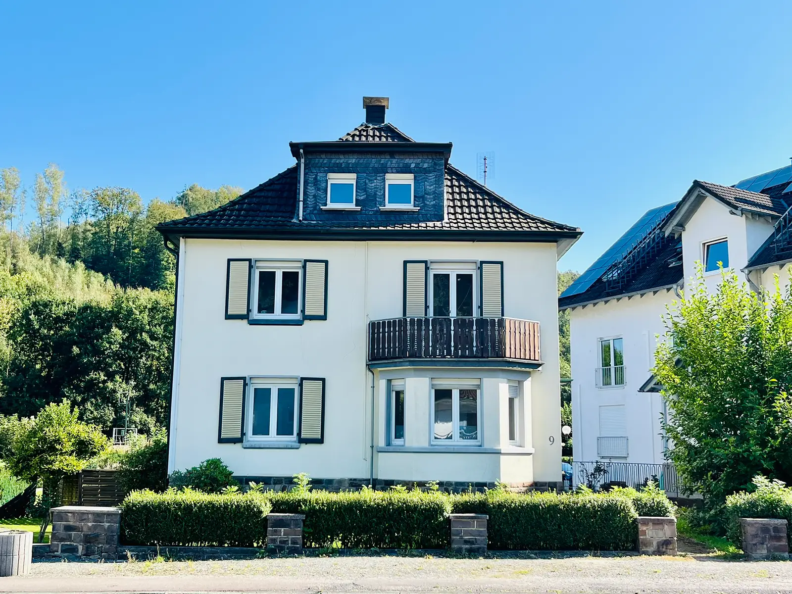 Objekt 2 Ein dreistöckiges weißes Haus mit einem dunklen Ziegeldach, Erkerfenstern und einem kleinen Balkon, umgeben von grünen Büschen und Bäumen unter einem klaren blauen Himmel.