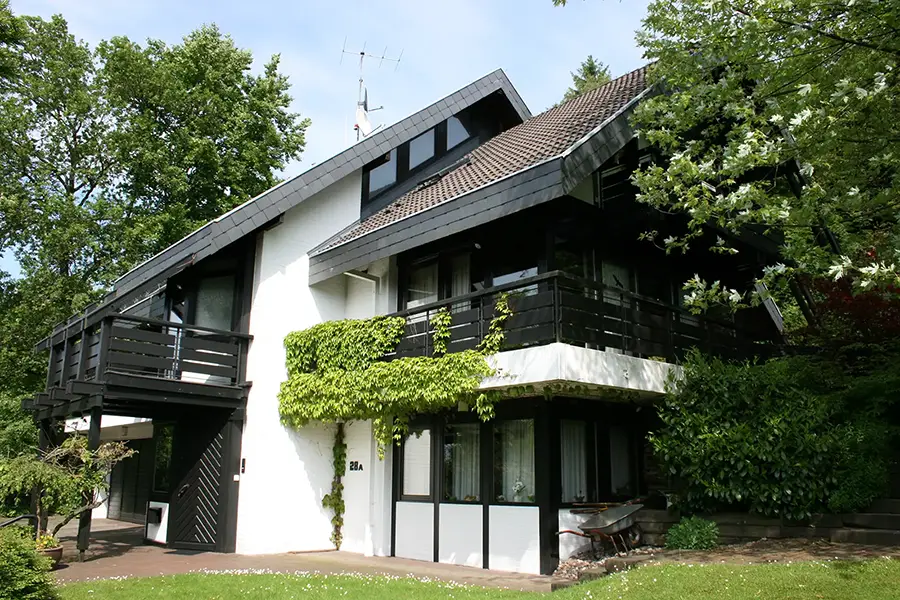 Objekt 20 Ein modernes zweistöckiges Haus mit weißen Wänden, dunklem Dach und schwarzem Geländer, umgeben von Grün und Bäumen, mit Efeu an der Fassade.