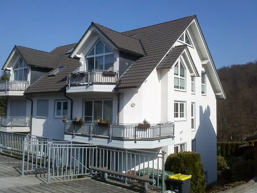 Objekt 21 Ein modernes, weißes, mehrstöckiges Haus mit großen Fenstern, Balkonen und einem schrägen, dunkelgrauen Dach, vor einem klaren, blauen Himmel und umgeben von Bäumen.