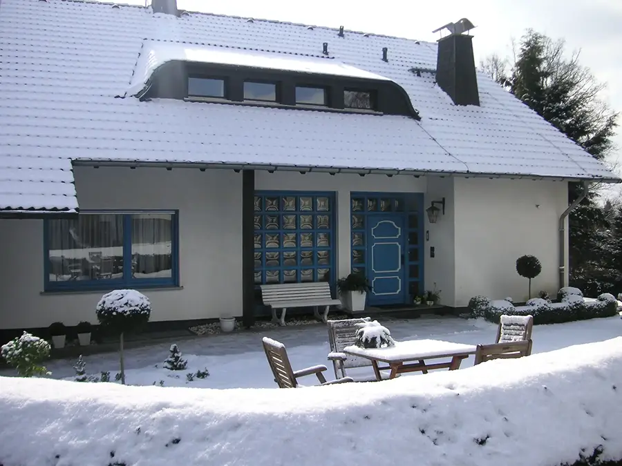 Objekt 22 Ein weißes Haus mit blauen Verzierungen steht im Schnee, mit einer blauen Tür, einem schneebedeckten Dach und einer Terrasse mit Gartenmöbeln.