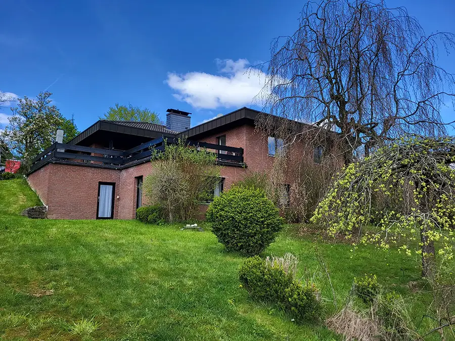 Objekt 23 Ein zweistöckiges Backsteinhaus mit einem Balkon, umgeben von grünem Gras, Büschen und Bäumen unter blauem Himmel.