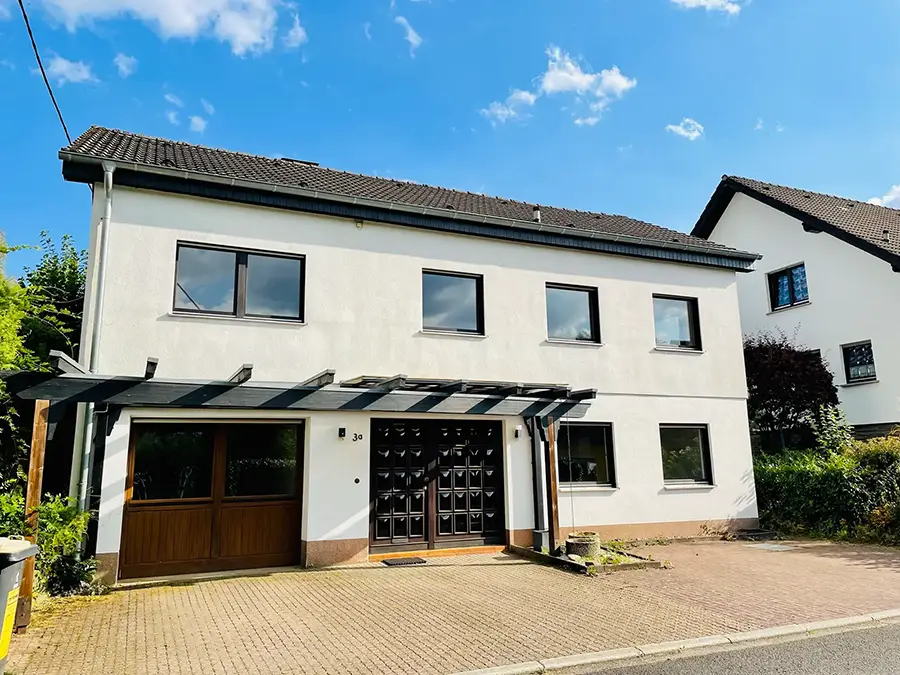 Objekt 24 Zweistöckiges weißes Haus mit dunklem Ziegeldach, schwarz umrahmten Fenstern und einem hölzernen Garagentor auf einer gepflasterten Auffahrt unter einem blauen Himmel mit vereinzelten Wolken.