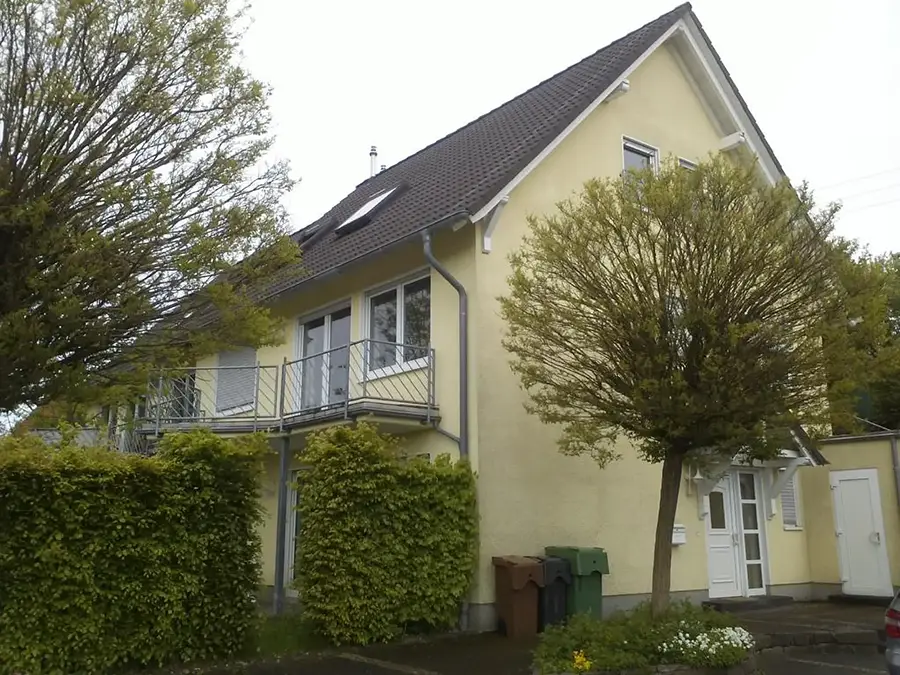 Objekt 25 Ein zweistöckiges gelbes Haus mit grauem Dach, Balkon und weiß gerahmten Fenstern, umgeben von Bäumen und Sträuchern mit mehreren Mülltonnen in der Nähe des Eingangs.