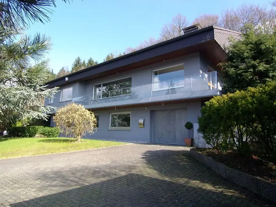 Objekt 26 Zweistöckiges Haus mit grauer Fassade, großen Fenstern, einem Balkon, einer Garage und einer Grünanlage vor dem Haus.