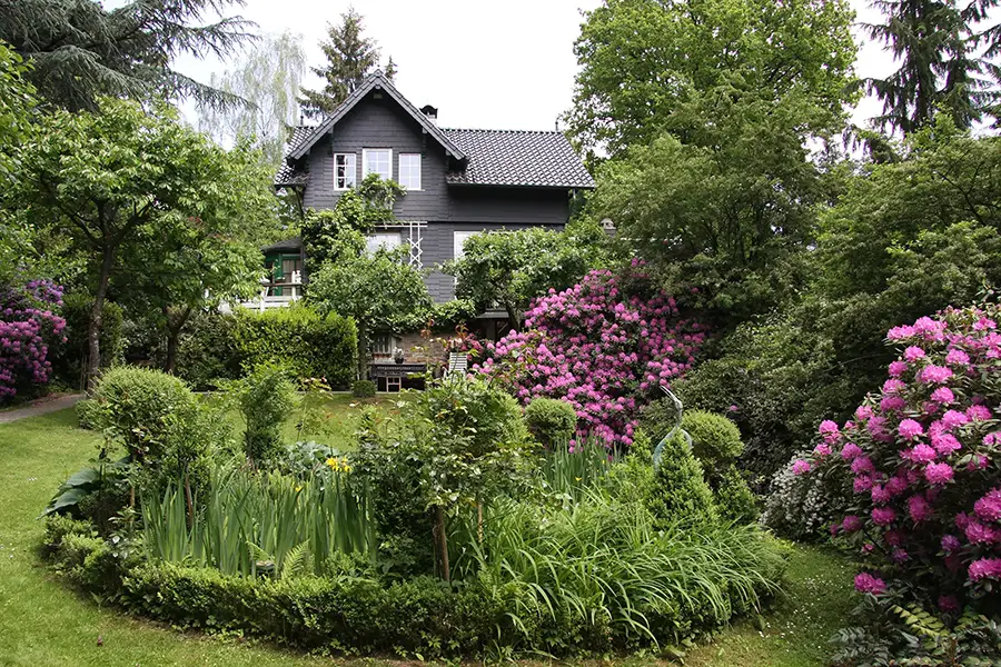 Objekt 27 Ein zweistöckiges dunkles Haus, umgeben von einem üppigen Garten mit grünen Sträuchern, hohen Bäumen und blühenden rosa und lila Blumen.