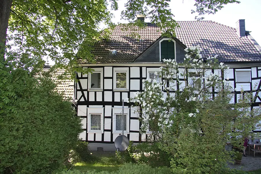 Objekt 28 Ein traditionelles Fachwerkhaus mit weißen Wänden und dunklen Holzbalken, umgeben von grünen Bäumen und Sträuchern, mit einer Satellitenschüssel an der Fassade.