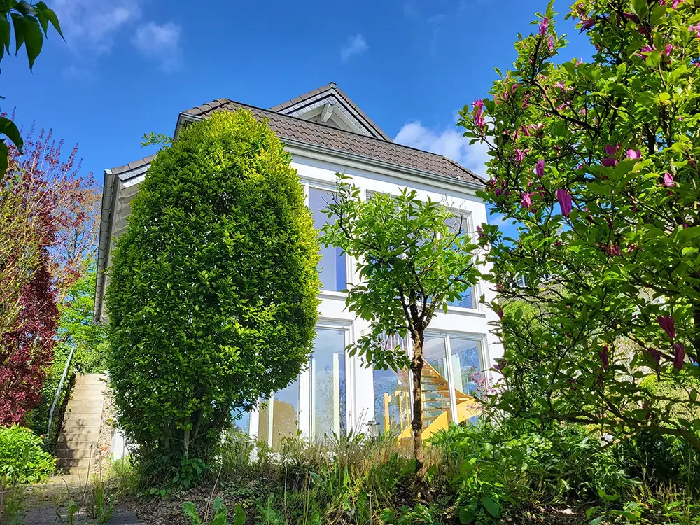 Objekt 30 Ein modernes weißes Haus mit großen Fenstern ist umgeben von grünen Bäumen und Sträuchern unter einem klaren blauen Himmel.