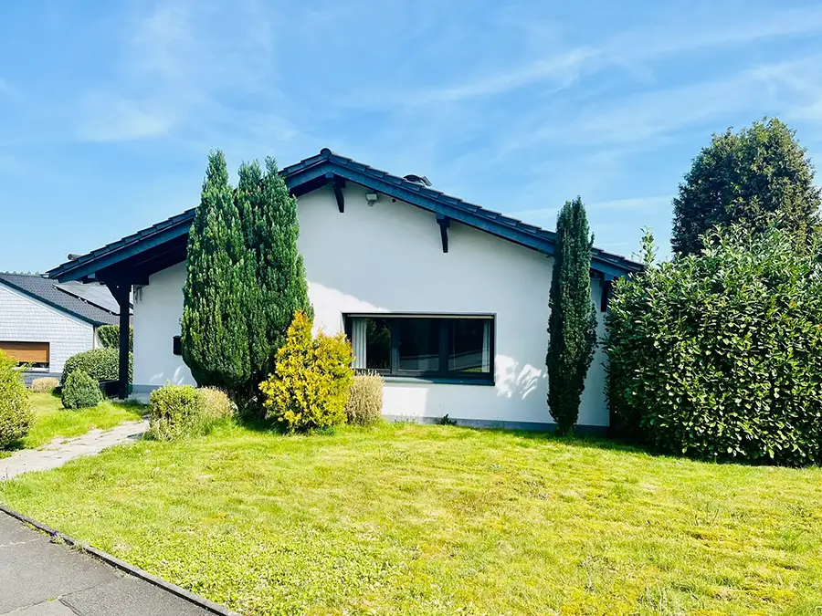Objekt 6 Ein einstöckiges weißes Haus mit einem dunklen Dach, einer großen Fensterfront und einem grünen Rasen mit verschiedenen Büschen und Bäumen unter einem klaren blauen Himmel.
