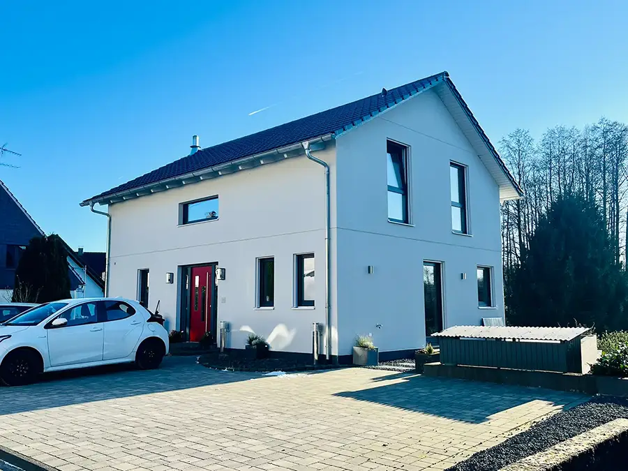 Objekt 7 Ein modernes zweistöckiges Haus mit weißen Außenwänden, einer roten Eingangstür und einem dunklen Ziegeldach, in dessen Einfahrt an einem sonnigen Tag ein weißes Auto parkt.